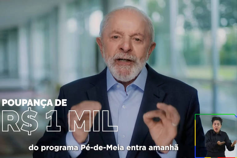 Um homem de cabelos brancos e barba， vestido com um paletó escuro e camisa clara， está falando em um ambiente iluminado e moderno. Ele gesticula com as mãos enquanto apresenta uma oferta de poupança de R$ 1 mil. À direita， há uma janela com uma pessoa fazendo interpretação em Libras. O texto na imagem diz: 039;POUPANÇA DE R$ 1 MIL do programa Pé-de-Meia entra amanhã039;.