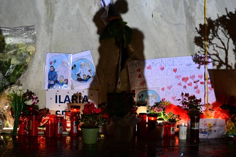 A imagem mostra um memorial com velas acesas e flores em frente a uma parede. Há cartazes com desenhos e mensagens， incluindo o nome 039;ILARIA039;. As flores estão dispostas em vasos e há uma variedade de velas， algumas em copos de vidro. A iluminação é suave， criando um ambiente de homenagem.