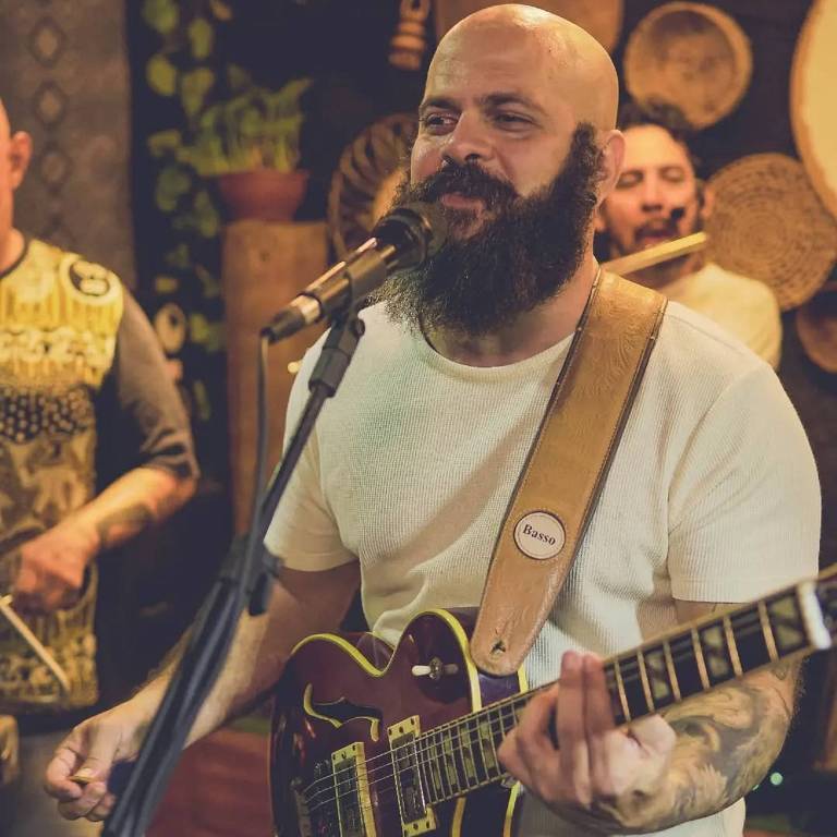 Um homem com barba e cabeça raspada está cantando em um microfone enquanto toca guitarra. Ele usa uma camiseta branca e tem uma correia de guitarra. Ao fundo， outros músicos podem ser vistos， e a decoração do ambiente inclui elementos rústicos e artesanais.