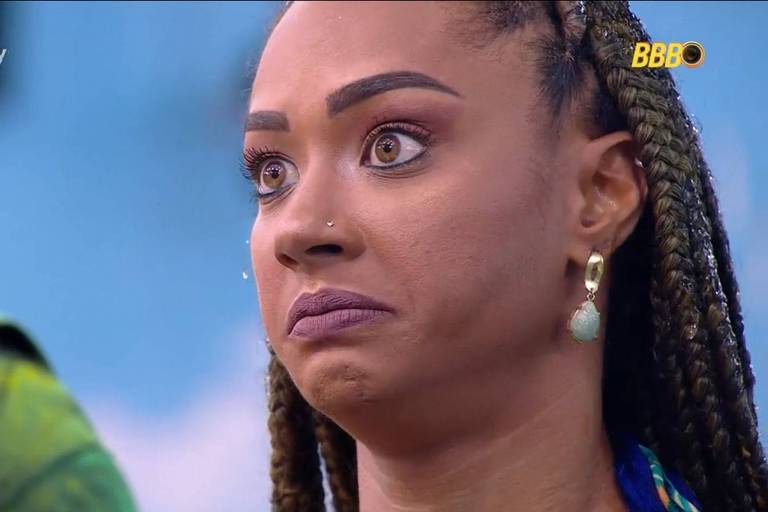 A imagem mostra uma mulher com cabelo trançado e brincos grandes, com uma expressão facial que transmite surpresa ou preocupação. Ela está olhando para a câmera, com os olhos bem abertos e a boca ligeiramente entreaberta. Ao fundo, há uma figura desfocada, possivelmente outra pessoa.