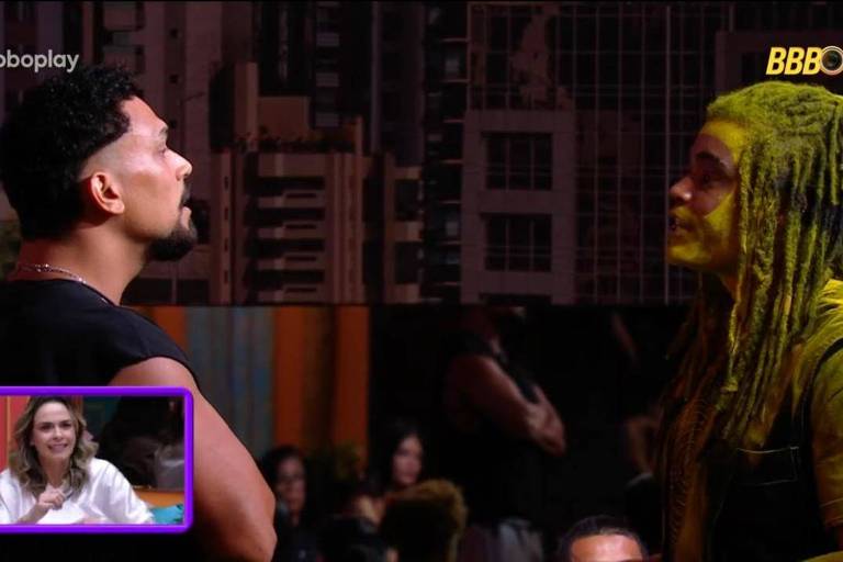 A imagem mostra um confronto entre dois homens. Um deles, à esquerda, tem cabelo curto e barba, enquanto o outro, à direita, tem cabelo em dreads e está pintado de amarelo. No canto inferior esquerdo, há uma mulher sentada em um sofá, observando a cena. Ao fundo, é possível ver uma parede com prédios e outras pessoas assistindo.