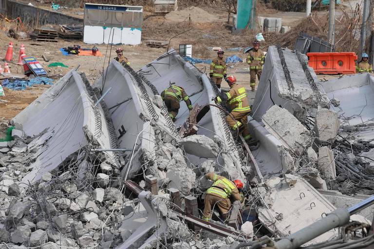 A imagem mostra bombeiros trabalhando em meio a escombros de uma estrutura colapsada. Eles estão cercados por destroços de concreto e aço， com alguns equipamentos de resgate visíveis ao fundo. O cenário é de destruição， com uma área ao redor que parece ter sido afetada por um desastre.