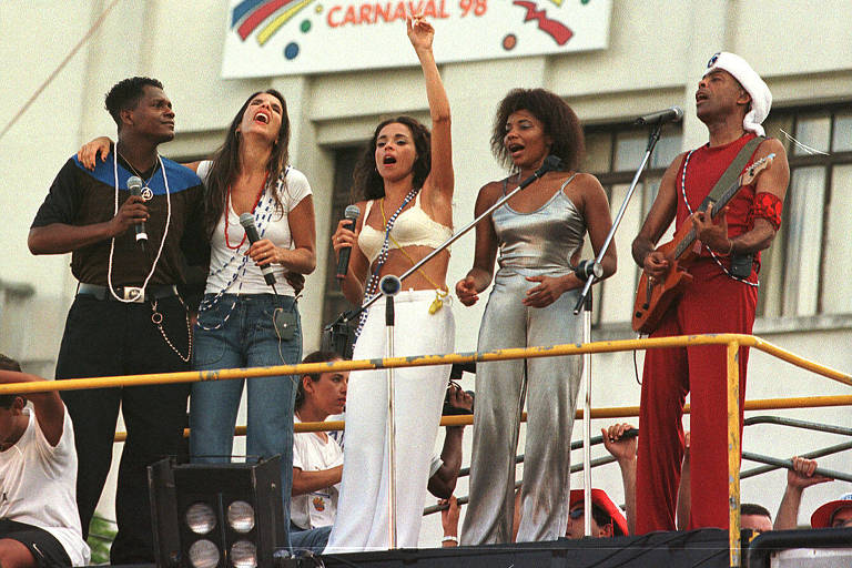 Cinco artistas estão se apresentando em um palco durante o Carnaval de 1998. Dois cantores estão segurando microfones, enquanto uma cantora levanta o braço em celebração. As mulheres estão vestidas com roupas brilhantes e justas, e um dos homens toca violão. Ao fundo, há uma faixa com a inscrição 'CARNAVAL 98'