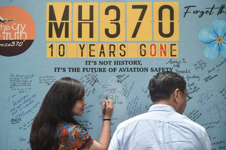 A imagem mostra um mural em homenagem ao desaparecimento do voo MH370， com a inscrição 039;MH370 10 YEARS GONE039; em letras grandes. Duas pessoas estão escrevendo mensagens no mural， que está coberto de assinaturas e mensagens de apoio. O mural também contém a frase 039;IT039;S NOT HISTORY， IT039;S THE FUTURE OF AVIATION SAFETY039;.