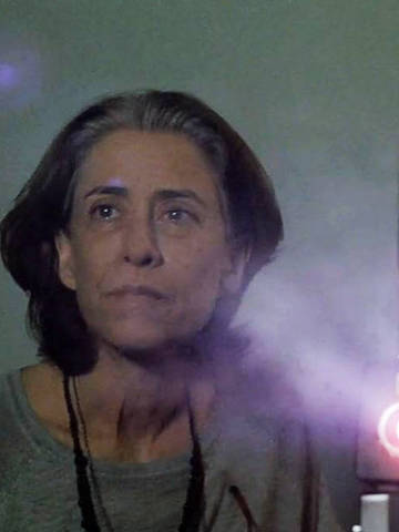 Cena do filme Ainda Estou Aqui *** Local Caption *** A imagem mostra uma mulher com cabelo escuro e grisalho, olhando atentamente para um projetor de filme. O projetor emite uma luz vermelha e há uma névoa visível ao redor, sugerindo que está em funcionamento. O fundo é de uma cor verde suave, e a mulher usa uma blusa cinza e colares. A expressão dela é séria e contemplativa.
