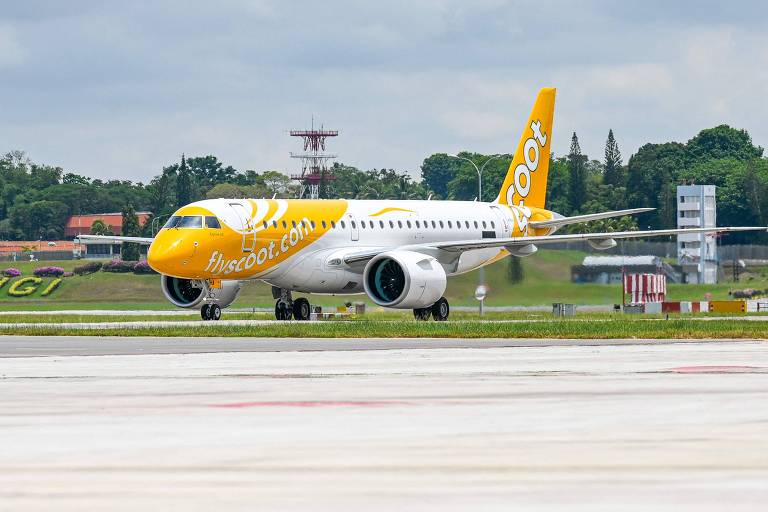 Um avião da companhia aérea Scoot， pintado de amarelo e branco， está taxiando na pista do Aeroporto de Changi. Ao fundo， há uma torre de controle e vegetação. O céu está nublado.