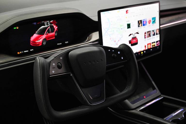 A imagem mostra o interior de um carro Tesla， com um volante moderno e uma tela central grande. O volante possui o logotipo da Tesla e botões de controle. À frente do volante， há um painel digital que exibe uma imagem de um carro vermelho. A tela central mostra uma interface com várias imagens e um mapa.