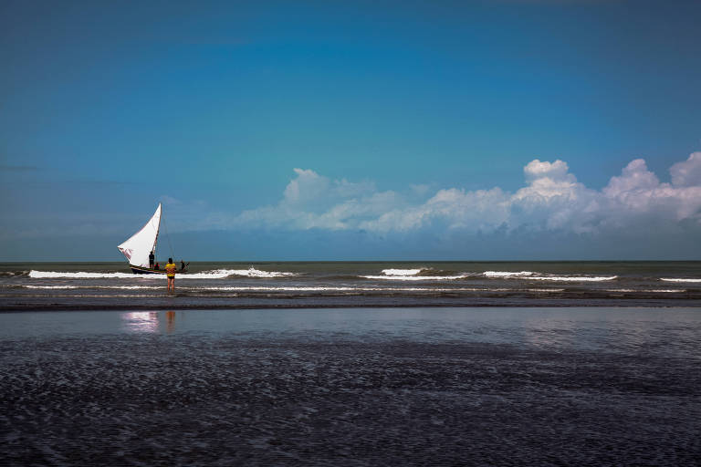 A imagem mostra uma praia com um barco à vela branco navegando ao fundo. Na areia, duas pessoas estão próximas à água, observando o mar. O céu é azul com algumas nuvens brancas, e as ondas do mar estão suaves.