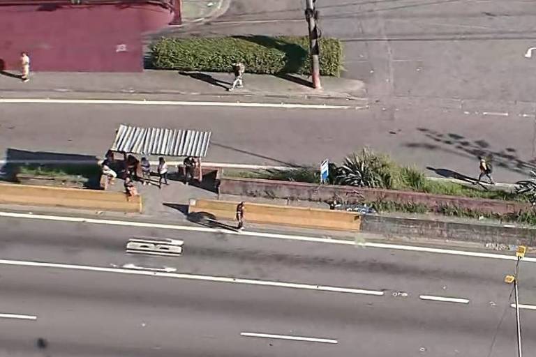 A imagem mostra uma parada de ônibus em uma área urbana. Há várias pessoas na calçada， algumas esperando na parada， que possui um telhado de metal. Ao fundo， é possível ver um trecho da estrada com faixas brancas e um canteiro com vegetação. O ambiente é ensolarado e há um muro vermelho ao fundo.