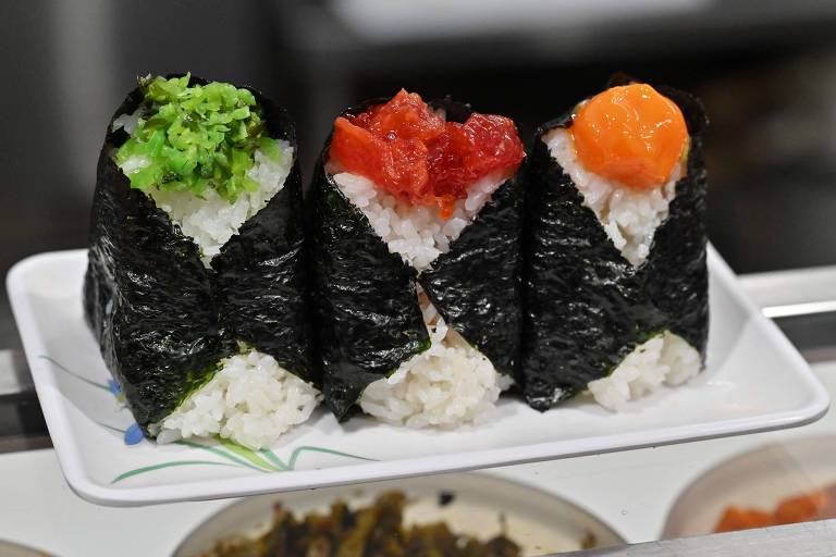 A imagem mostra três cones dispostos em um prato branco. Cada cone é envolto em alga nori e contém arroz. O cone da esquerda é coberto com folhas verdes， o do meio tem uma camada de atum vermelho， e o da direita apresenta uma cobertura laranja， possivelmente de ovas. Ao fundo， há outros pratos com acompanhamentos.