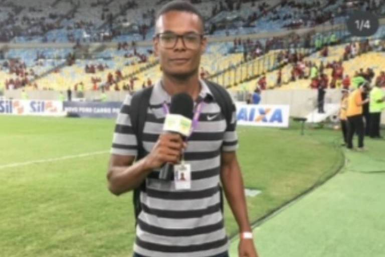 Um repórter está em um campo de futebol， segurando um microfone e usando óculos. Ele veste uma camisa listrada em preto e branco e calças escuras. Ao fundo， há uma arquibancada com torcedores e bandeiras， e o gramado é visível.