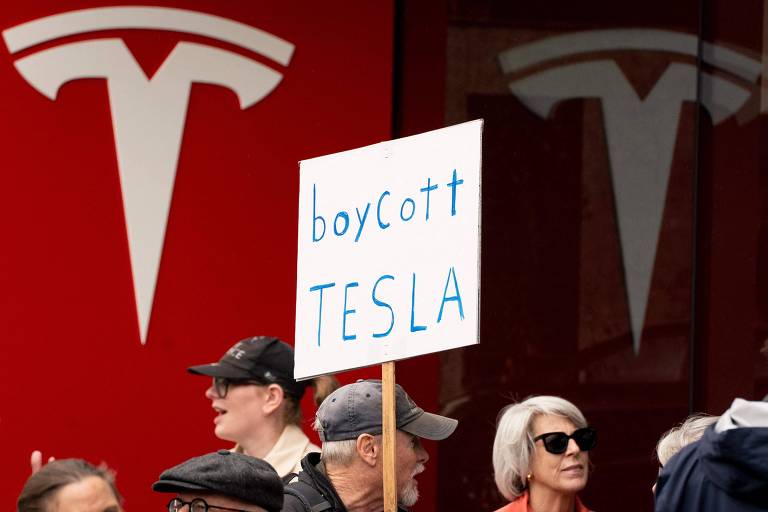 A imagem mostra um grupo de pessoas em um protesto， com um foco em um cartaz que diz 039;boycott TESLA039;. Ao fundo， há o logotipo da Tesla em uma parede vermelha. Algumas pessoas estão usando óculos escuros e chapéus， enquanto outras observam o protesto.