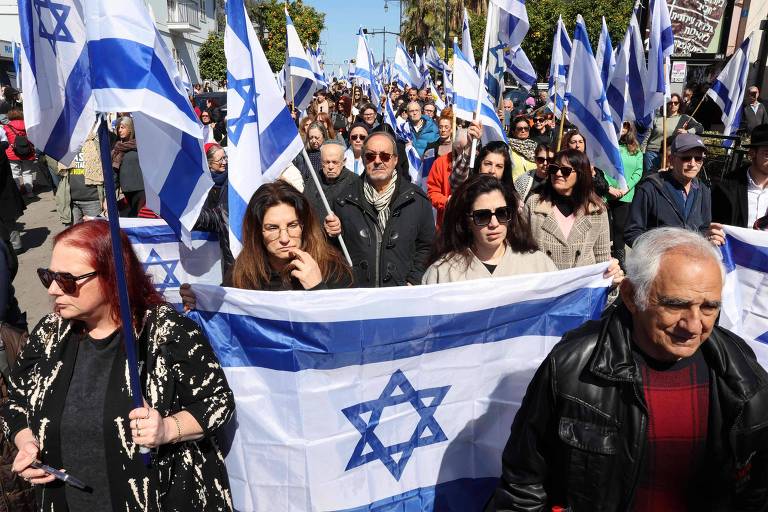 A imagem mostra uma grande manifestação com várias pessoas segurando bandeiras de Israel. O grupo é composto por homens e mulheres de diferentes idades， alguns usando óculos e outros com cabelos de diferentes cores. No centro， uma mulher segura uma bandeira maior， enquanto outras pessoas ao redor também seguram bandeiras menores. O ambiente parece ser ao ar livre， com árvores e um céu claro ao fundo.