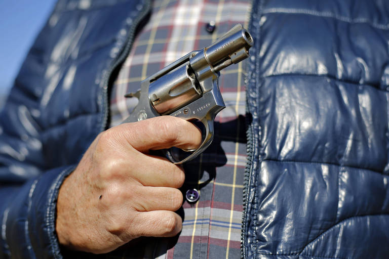 A imagem mostra uma mão segurando um revólver. A pessoa está vestindo uma jaqueta acolchoada azul e uma camisa xadrez. O fundo é desfocado， mas parece ser um ambiente ao ar livre.