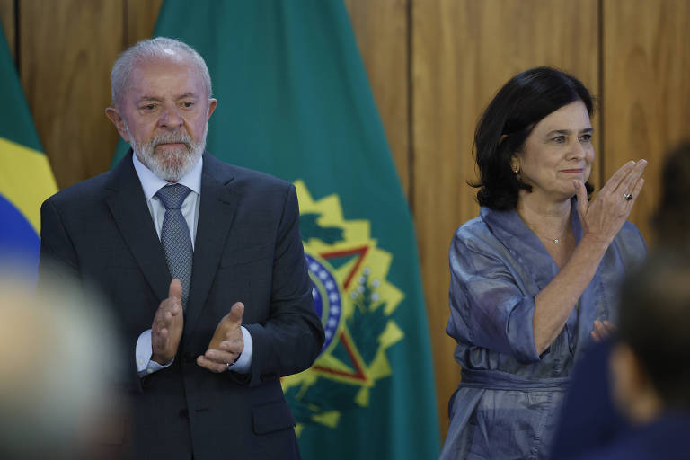 Imagem de uma cerimônia oficial com duas pessoas em destaque. À esquerda， um homem de cabelo grisalho e barba， vestindo um terno escuro e gravata， aplaudindo. À direita， uma mulher com cabelo escuro e liso， usando uma blusa azul， também aplaudindo. Ao fundo， bandeiras do Brasil e um símbolo nacional.