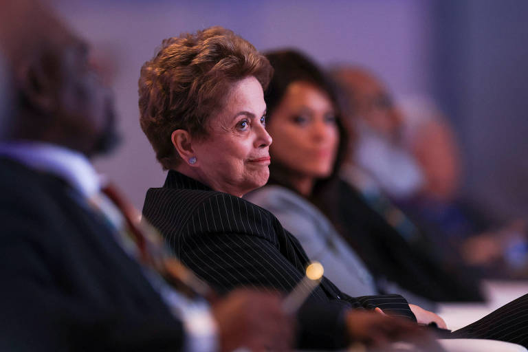 A imagem mostra um grupo de pessoas assistindo a um evento. Em primeiro plano， uma mulher de cabelo curto e cacheado， vestindo um terno listrado， está olhando para o lado. Ao fundo， há outras duas pessoas， uma delas com cabelo liso e escuro. O ambiente parece ser uma conferência ou seminário， com iluminação suave.