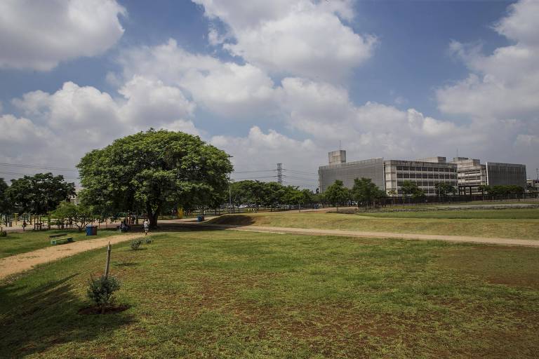 A imagem mostra um parque amplo com um gramado verde e algumas árvores. No fundo, há um edifício moderno de vários andares. O céu está parcialmente nublado, com nuvens brancas e um fundo azul. Há também um caminho de terra que atravessa o parque.