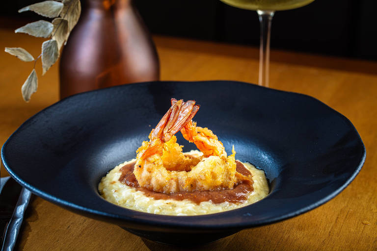 Um prato de risoto servido em um prato preto. O risoto é cremoso e possui um molho marrom por cima. Dois camarões empanados e fritos estão decorando o prato, posicionados de forma a parecerem em pé. Ao fundo, há uma garrafa de vidro e um copo com bebida.