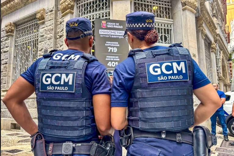 A imagem mostra dois guardas civis metropolitanos de costas， usando coletes táticos pretos com as inscrições 039;GCM039; e 039;SÃO PAULO039;. Eles estão em frente a um prédio， observando um cartaz que contém informações sobre um serviço público. O ambiente é urbano， com detalhes arquitetônicos visíveis ao fundo.