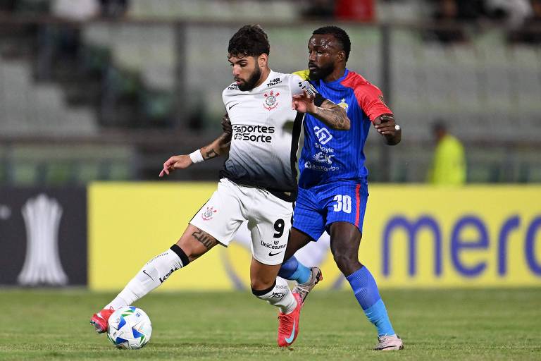 A imagem mostra dois jogadores de futebol em um campo. O jogador à esquerda， vestindo uma camisa branca com detalhes em preto e o número 9， está em movimento， enquanto o jogador à direita， com uma camisa azul e detalhes vermelhos， está tentando desarmá-lo. Ao fundo， há uma arquibancada e um banner publicitário.