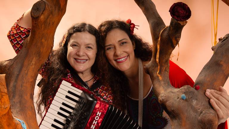 A imagem mostra duas mulheres sorrindo， uma segurando um acordeão. Elas estão posicionadas entre galhos de uma árvore， com um fundo de cor clara. Ambas têm cabelos longos e soltos， e estão vestidas com roupas coloridas. A mulher à esquerda usa uma blusa com padrões e a outra está com uma blusa mais simples. Ambas têm flores no cabelo.