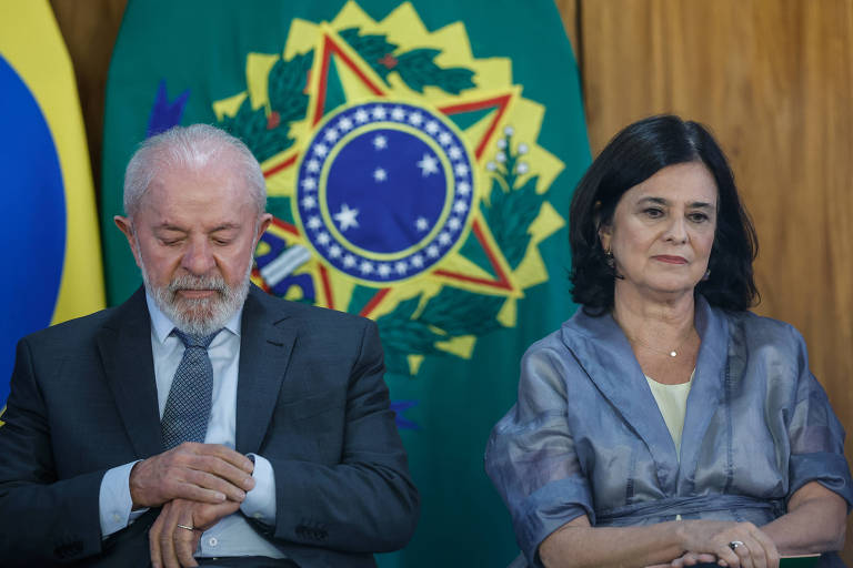 A imagem mostra duas pessoas sentadas em um evento oficial. À esquerda， um homem com cabelo grisalho e barba， vestindo um terno escuro e gravata， está com a cabeça baixa e as mãos cruzadas. À direita， uma mulher com cabelo escuro e liso， vestindo uma blusa cinza， está olhando para frente com uma expressão séria. Ao fundo， há bandeiras do Brasil e um grande símbolo nacional.