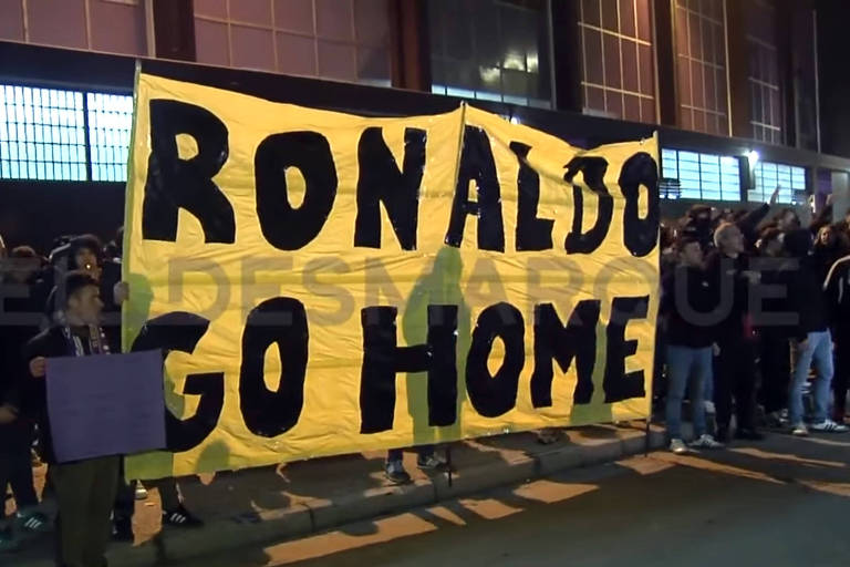 A imagem mostra um grupo de pessoas reunidas em um protesto à noite. Eles seguram uma grande faixa amarela com as palavras 039;RONALDO GO HOME039; escritas em letras pretas. O ambiente parece ser urbano， com edifícios ao fundo e iluminação artificial.