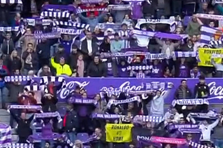 A imagem mostra um estádio de futebol com torcedores segurando faixas e cachecóis em tons de roxo e branco. Jogadores em uniformes verdes e laranjas estão se preparando para o jogo， enquanto crianças vestindo camisetas brancas caminham ao lado deles. O ambiente é festivo， com muitos torcedores ao fundo.