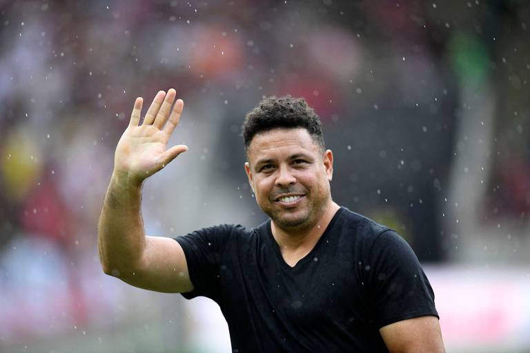 Um homem com cabelo curto e ondulado， vestindo uma camiseta preta， está acenando com a mão direita. Ele sorri e parece estar em um ambiente ao ar livre， com uma leve chuva visível ao fundo. O cenário é desfocado， mas há pessoas e cores que sugerem um evento esportivo.