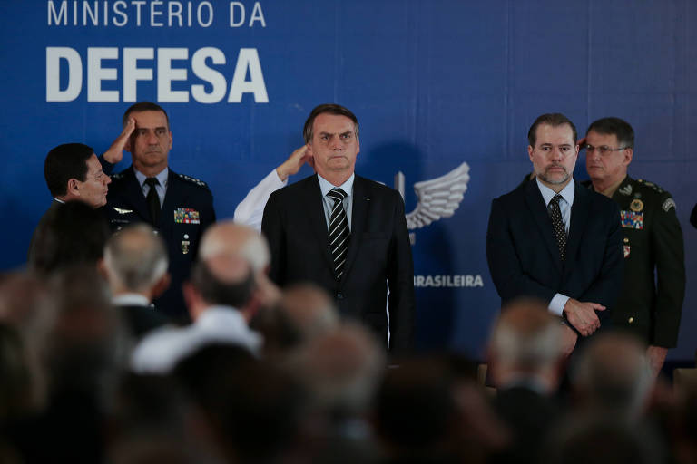 A imagem mostra uma cerimônia no Ministério da Defesa do Brasil. No centro， um homem em terno escuro está em pé， com uma expressão séria. Ao seu redor， outras pessoas， incluindo militares em uniformes， estão em posição de saudação. Ao fundo， uma parede azul com a inscrição 039;MINISTÉRIO DA DEFESA039;.