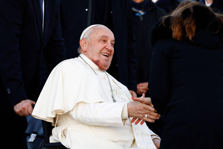 Imagem do Papa Francisco sorrindo e estendendo a mão para cumprimentar uma pessoa， enquanto está sentado em uma cadeira de rodas. Ele está vestido com uma batina branca e um manto， cercado por outras pessoas em um ambiente ao ar livre.