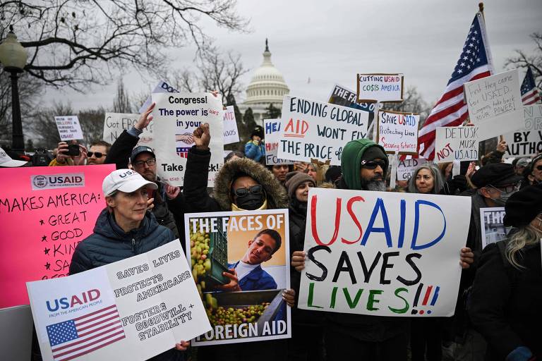 A imagem mostra um grupo de manifestantes em frente ao Capitólio dos Estados Unidos， segurando cartazes e bandeiras. Os cartazes incluem mensagens como 039;USAID Saves Lives!!!039; e 039;Ending Global Aid Won039;t Fix the Price039;. Os manifestantes estão vestidos com roupas de inverno， alguns usando bonés e cachecóis. O ambiente é de protesto， com várias pessoas visíveis e árvores ao fundo.