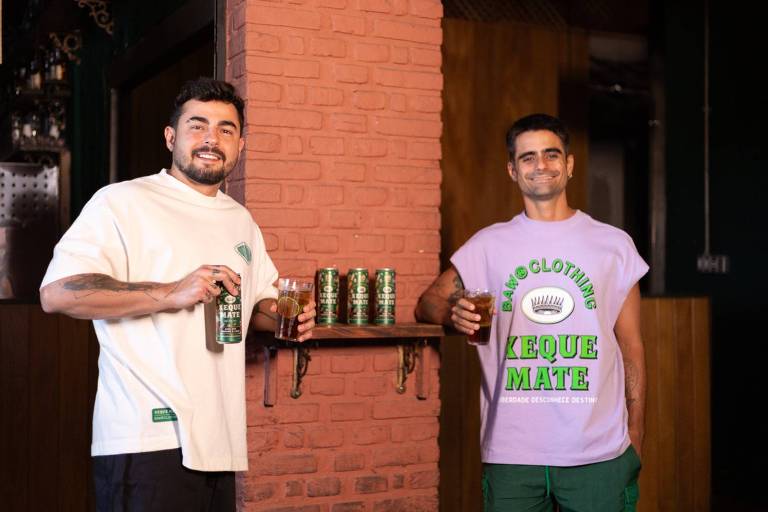 Dois homens estão em um ambiente interno， sorrindo e segurando latas de bebida. O homem à esquerda usa uma camiseta branca e segura uma lata na mão， enquanto o homem à direita veste uma camiseta lilás com a inscrição 039;XEQUE MATE039; e também segura uma lata. Ao fundo， há uma prateleira com várias latas de bebida dispostas.