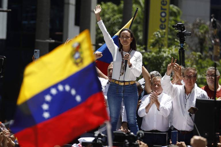 A imagem mostra uma mulher em pé em um palco， levantando a mão em sinal de saudação ou vitória. Ela está vestida com uma camisa branca e calças jeans. Ao fundo， há uma bandeira da Venezuela sendo agitada. Ao lado dela， algumas pessoas aplaudem， e há câmeras e equipamentos de gravação. O ambiente parece ser um evento ao ar livre， com árvores e edifícios ao fundo.