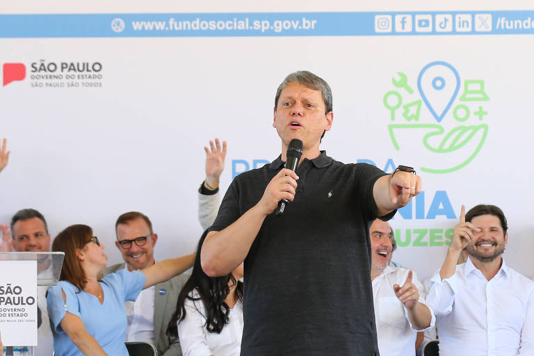 A imagem mostra um evento público em São Paulo， onde um homem está em pé， falando ao microfone. Ele usa uma camiseta escura e calças jeans. Atrás dele， há um painel com o logo da Fundação Social e outras informações. Várias pessoas estão sentadas em uma fila， algumas levantando as mãos. À frente， um grupo de pessoas observa e tira fotos. O ambiente parece ser ao ar livre， com uma estrutura montada para o evento.