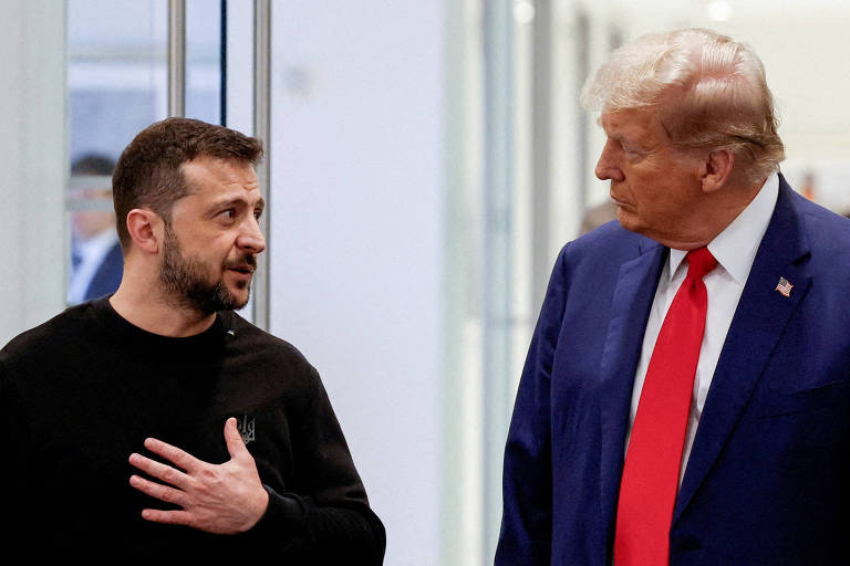 A imagem mostra dois homens conversando em um ambiente interno. Um deles está vestindo uma camiseta preta e gesticula com a mão esquerda， enquanto o outro usa um terno azul e uma gravata vermelha. O fundo é composto por paredes claras e portas de vidro.