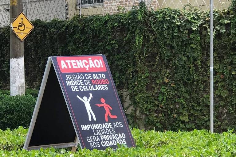 Uma placa de aviso em uma área urbana， destacando que é uma região de alto índice de roubo de celulares. A placa é preta com letras vermelhas e brancas， e ilustra uma figura humana em uma posição que sugere um ato de roubo. Ao fundo， há uma cerca verde e placas de rua indicando os nomes 039;Macaé039; e 039;Batalha039;.