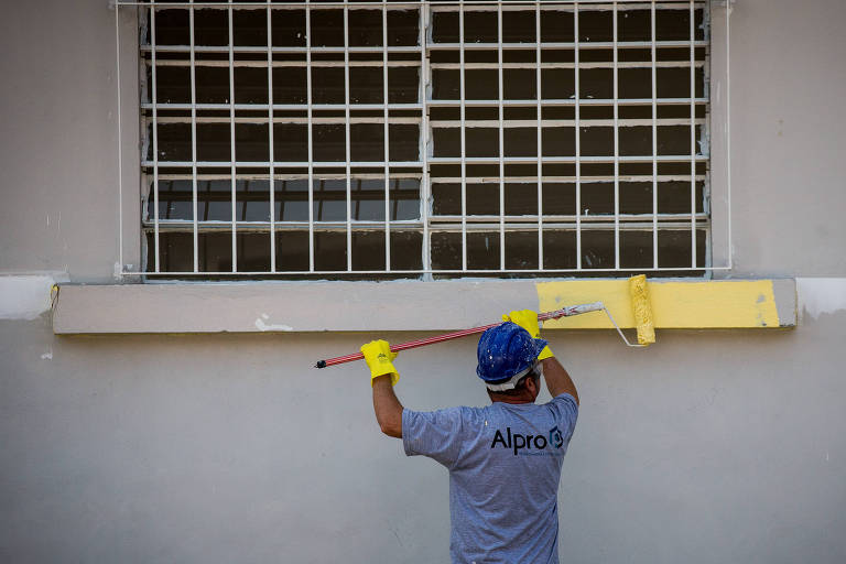 Um trabalhador está pintando uma parede. Ele usa uma camiseta cinza com a marca 039;Alpro039;， luvas amarelas e um capacete azul. O trabalhador está de costas， utilizando uma ferramenta longa para aplicar tinta na parte superior da parede. Ao fundo， há uma janela com grades e a parede está parcialmente pintada.