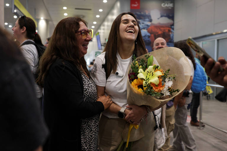 A imagem mostra duas mulheres sorrindo e se abraçando em um aeroporto. Uma delas segura um grande buquê de flores coloridas， enquanto a outra parece estar muito feliz. Ao fundo， há outras pessoas e uma decoração típica de aeroporto.