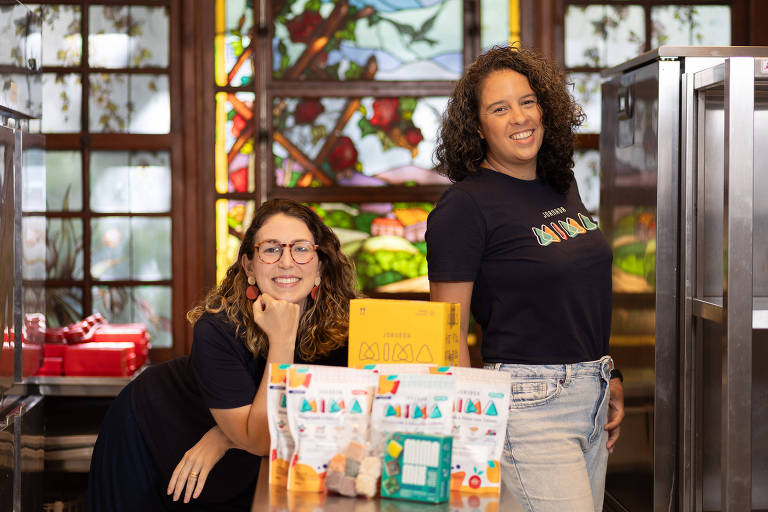 A imagem mostra duas mulheres sorrindo em um ambiente de cozinha. A mulher à esquerda está com os braços cruzados e apoiada em uma mesa， usando óculos e uma camiseta preta. A mulher à direita está em pé， tem o cabelo cacheado e também vestindo uma camiseta preta. Na mesa à frente delas， há produtos embalados e uma caixa amarela. Ao fundo， há janelas com vitrais coloridos.