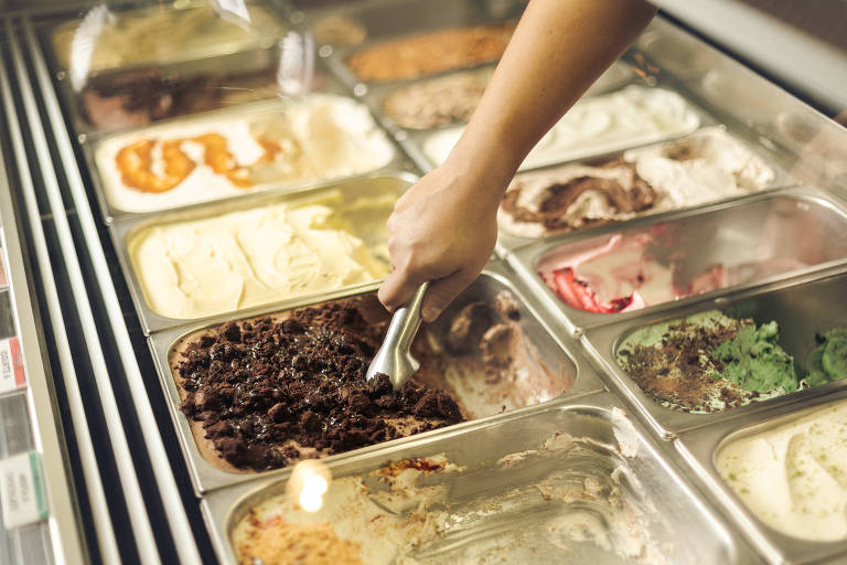 A imagem mostra um balcão de sorveteria com várias bandejas de sorvete. Uma mão está usando uma colher para servir o sorvete de chocolate， que está na bandeja inferior esquerda. As bandejas contêm diferentes sabores de sorvete， incluindo opções claras e escuras， com texturas variadas.