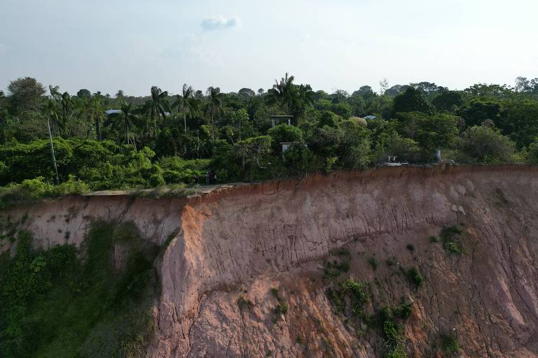 A imagem mostra um deslizamento de terra em uma área verde. A parte superior da imagem apresenta uma vegetação densa， com árvores e arbustos. A parte inferior revela um desfiladeiro com uma encosta exposta， onde o solo é de cor avermelhada e apresenta marcas de erosão. O céu está claro， com algumas nuvens visíveis.