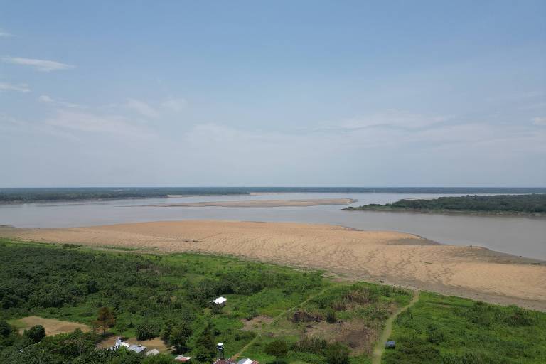 A imagem mostra uma vista panorâmica de um rio com áreas de areia exposta e vegetação ao redor. O céu está claro， com algumas nuvens， e a água do rio apresenta uma coloração marrom. Na margem do rio， há uma vegetação densa e algumas construções pequenas visíveis.