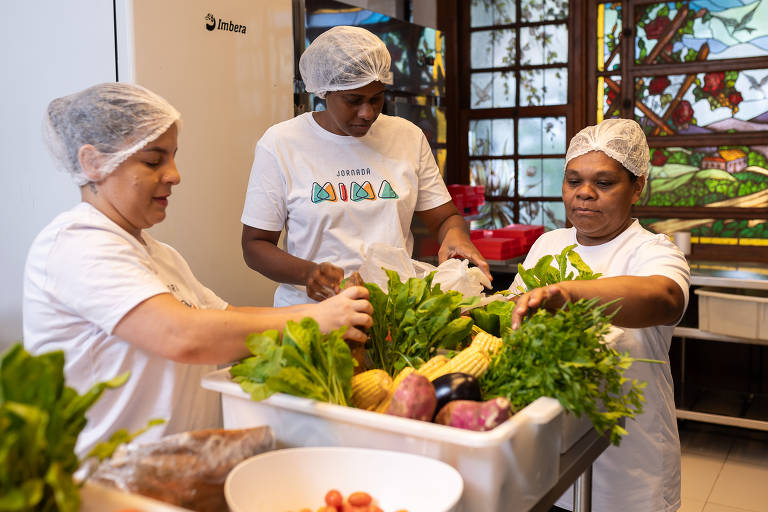 Três mulheres estão trabalhando em uma cozinha, organizando e preparando vegetais frescos. Elas usam toucas brancas e camisetas de cores claras. Uma mulher à esquerda segura um maço de folhas verdes, enquanto outra mulher no centro está colocando vegetais em uma caixa. A terceira mulher à direita também está manuseando vegetais. Ao fundo, há uma janela com vitrais coloridos.