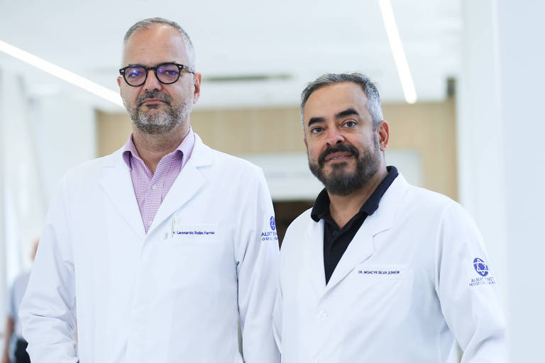 A imagem mostra dois homens vestidos com jalecos brancos de médicos. O primeiro homem， à esquerda， tem cabelo curto e grisalho， usa óculos e uma camisa rosa por baixo do jaleco. O segundo homem， à direita， tem cabelo escuro e barba， e está usando uma camisa preta sob o jaleco. Eles estão em um ambiente clínico moderno， com paredes claras e iluminação natural.