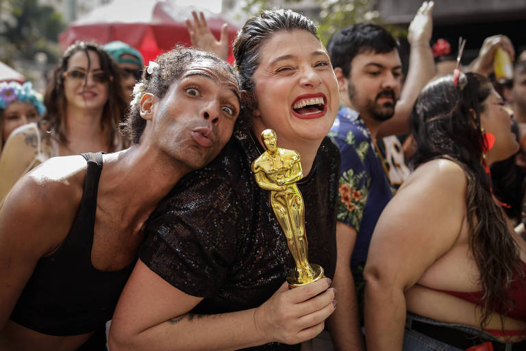 A imagem mostra um grupo de pessoas em um ambiente festivo， possivelmente durante um carnaval. Em primeiro plano， duas pessoas estatilde;o sorrindo. Uma delas， agrave; esquerda， tem cabelo cacheado e usa uma blusa preta， enquanto a outra， agrave; direita， tem cabelo liso e usa uma blusa preta com detalhes. Ela segura uma estatueta dourada， semelhante ao Oscar. Ao fundo， haacute; vaacute;rias outras pessoas， algumas com roupas coloridas e acessoacute;rios festivos.