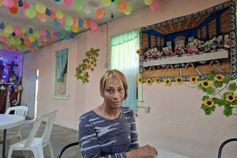 A imagem mostra uma mulher sentada em uma mesa em um ambiente decorado. O teto está coberto com balões coloridos， e as paredes têm uma pintura que representa a Última Ceia， além de elementos decorativos como girassóis e folhas verdes. A mulher tem cabelo curto e liso， e usa uma blusa listrada em tons de cinza. Ao fundo， há janelas com cortinas claras e mesas brancas.