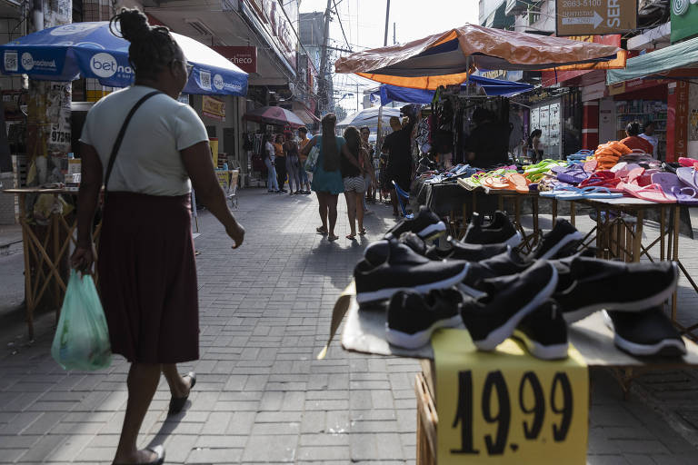 A imagem mostra uma rua movimentada com barracas de feira. À esquerda， uma mulher caminha carregando uma sacola. À direita， há uma mesa com sapatos pretos expostos， com um preço de R$ 19，99. Ao fundo， outras pessoas estão caminhando e há barracas cobertas com toldos coloridos.