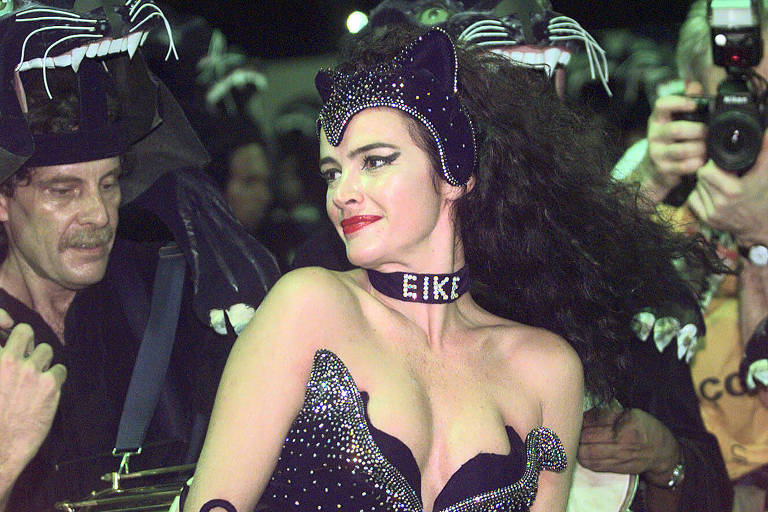 A imagem mostra uma mulher com uma fantasia brilhante de pantera, adornada com lantejoulas. Ela usa um colar com o nome 'EIKE' e uma tiara com orelhas de pantera. Ao fundo, há homens com fantasias de pantera e um fotógrafo capturando o momento. A cena parece ser de um evento de carnaval.