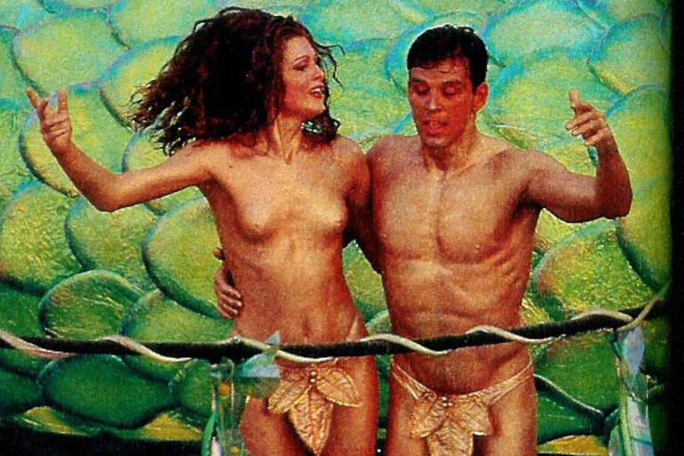 A imagem mostra um casal nu, posando em um ambiente que simula o fundo do mar. Eles estão em uma posição descontraída, com a mulher à esquerda e o homem à direita. Ambos usam acessórios que lembram escamas ou conchas na parte inferior do corpo. O fundo é composto por grandes pedras verdes, que imitam algas ou rochas subaquáticas.